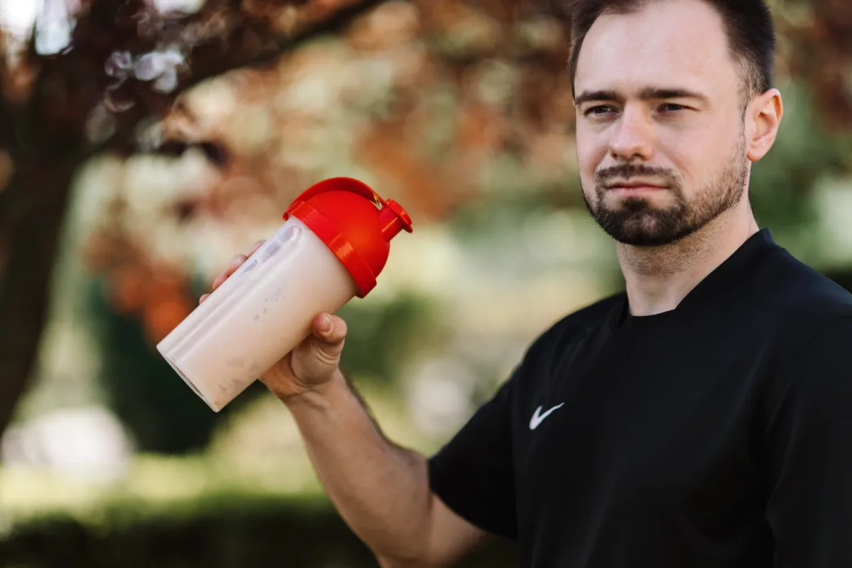 Man holding a protein shake tumbler outdoors in a black shirt