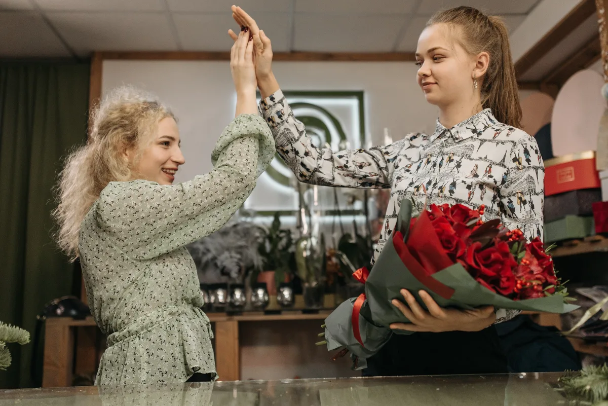 Two women laughing and exchanging red flowers indoors