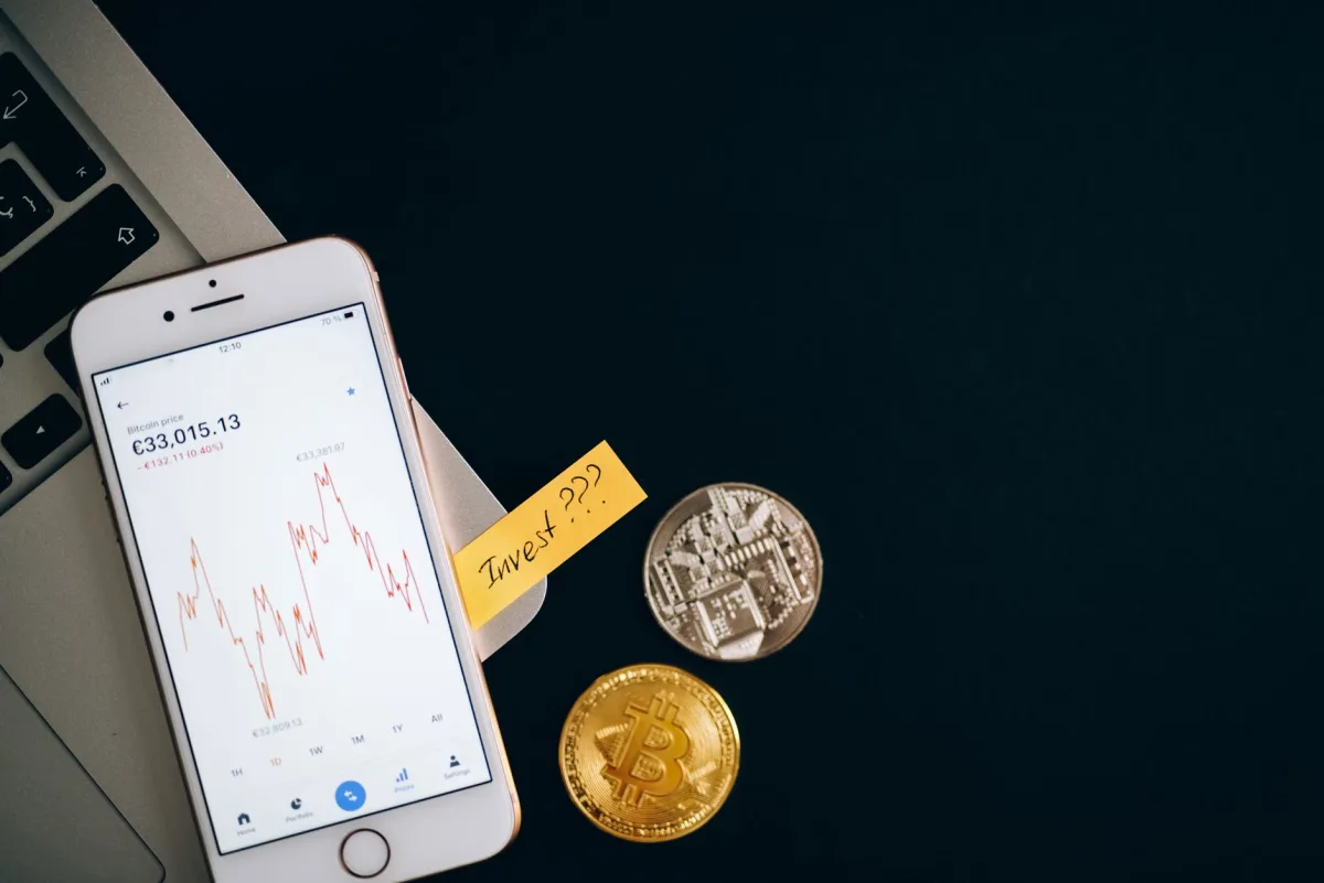 Smartphone displaying cryptocurrency prices beside coins and laptop