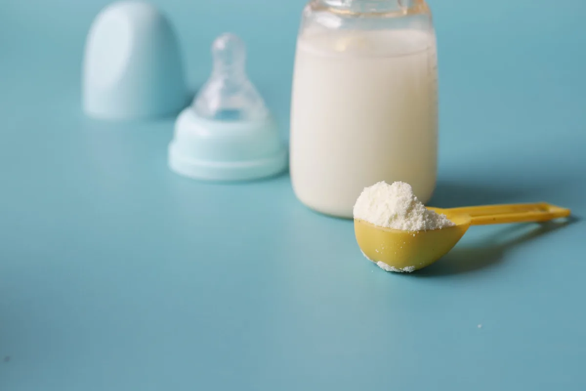 Baby formula powder in a measuring scoop next to a feeding bottle on a blue background