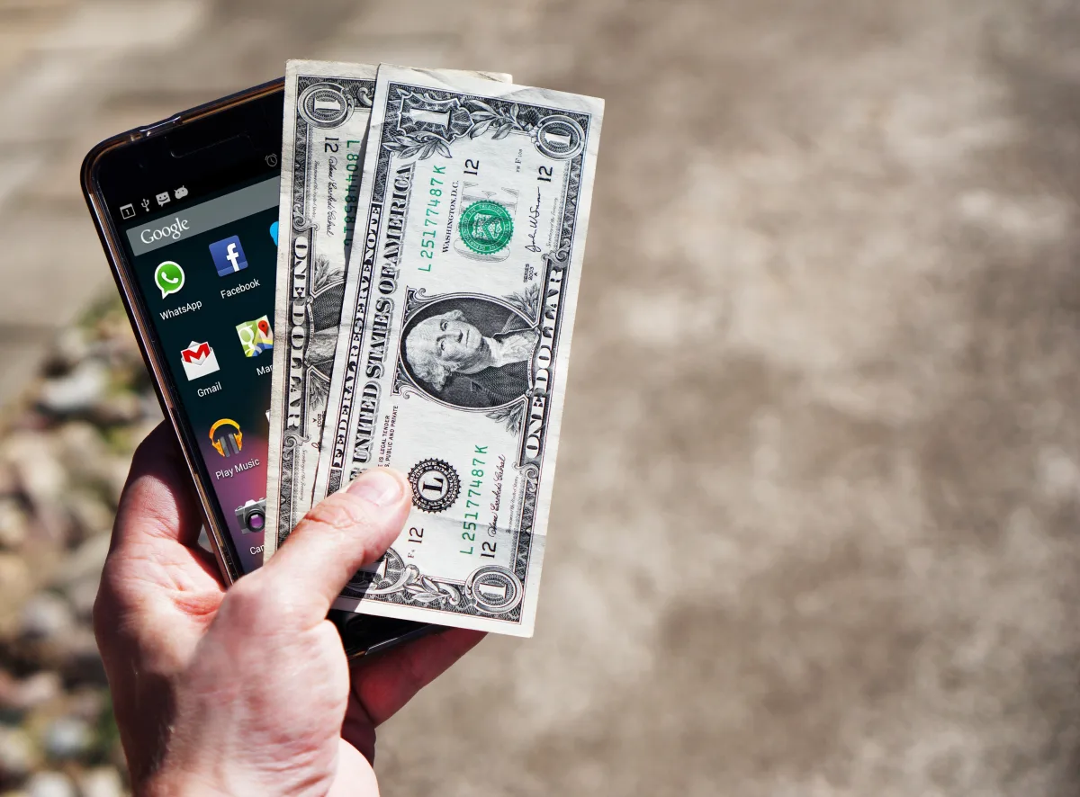 Hand holding US dollar bills and smartphone outdoors