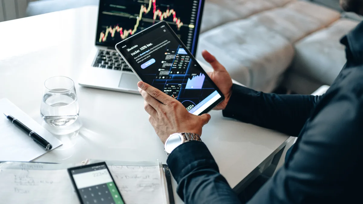 Person holding a tablet showing investment charts beside a laptop and notebook