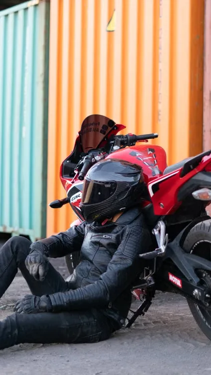 Biker wearing leather jacket and motorcycle helmet sitting next to a red sport bike