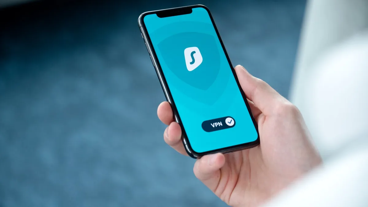 Hand holding smartphone with a VPN app open, laptop visible in the background