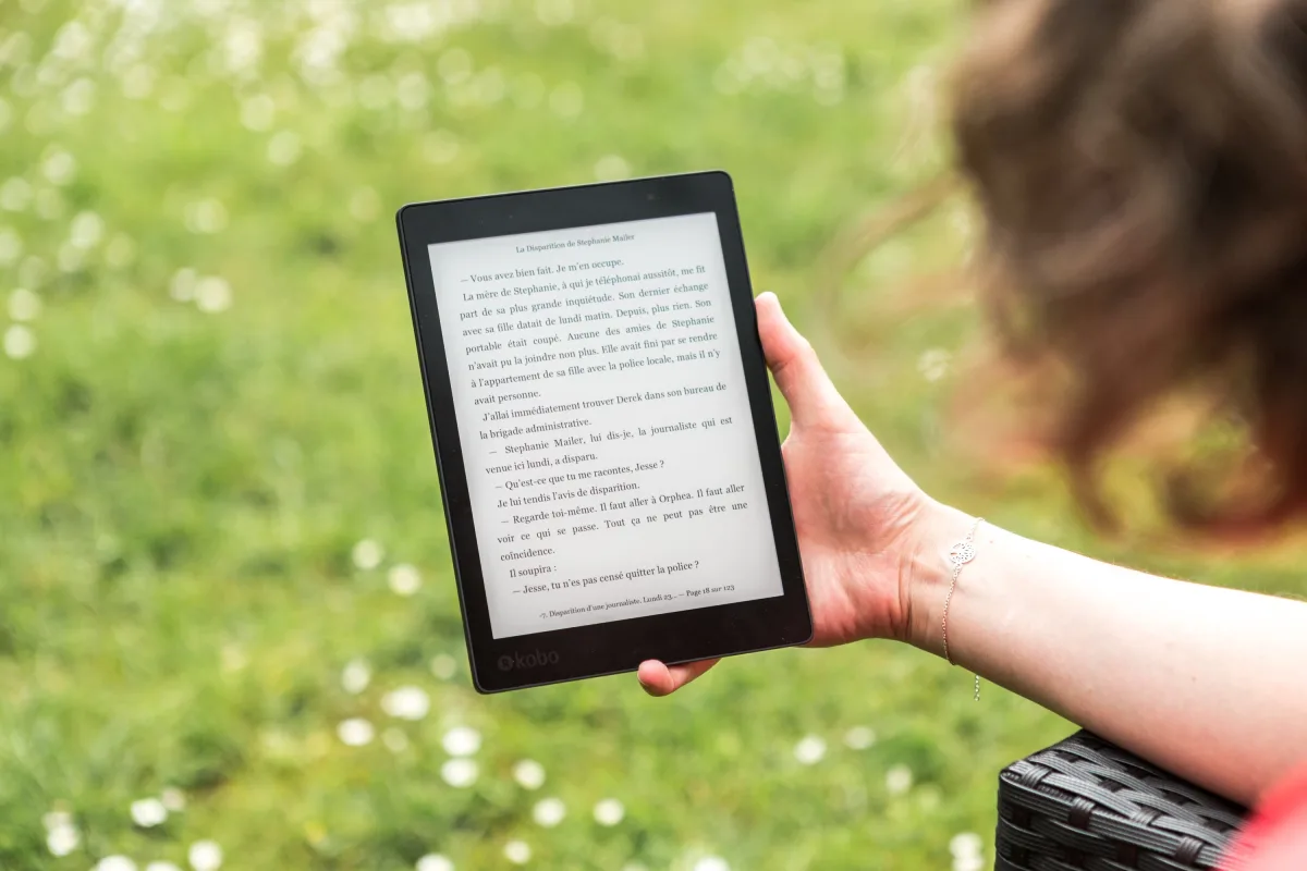 A person reads an eBook on a Kobo device while relaxing outdoors in a sunny park