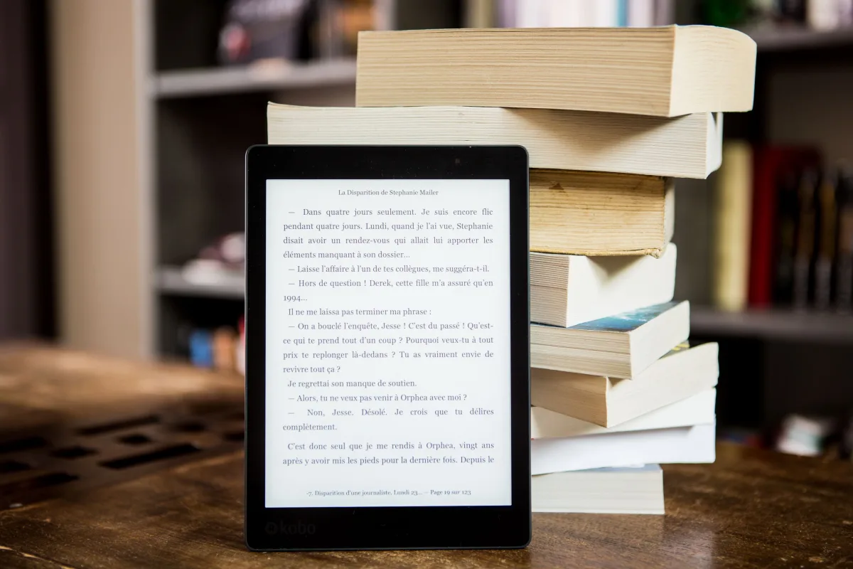 E-reader with text displayed next to a stack of physical books on a wooden table
