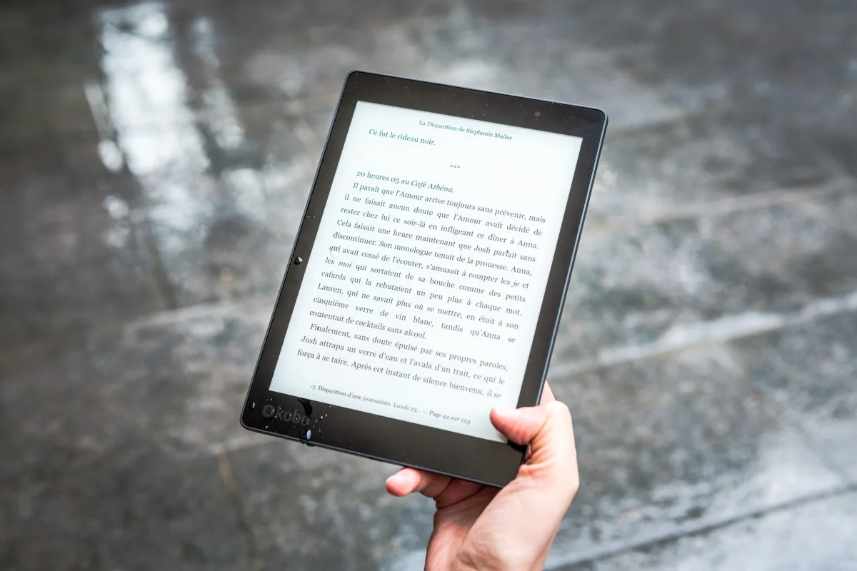 A person holds an e-reader displaying text against a wet pavement background