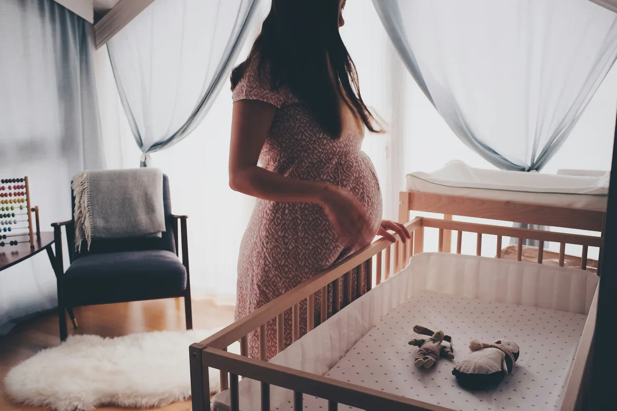 Pregnant woman standing beside a natural brown wooden crib in a bright nursery