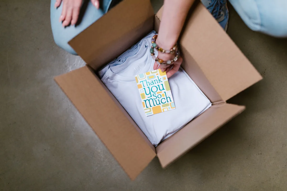 Person placing a folded white shirt into a cardboard shipping box with a thank-you card