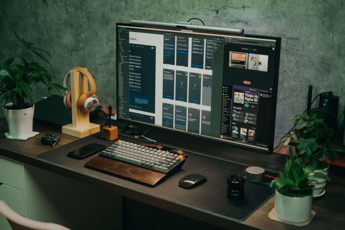 Home office desk with large monitor, keyboard, headphones, and plants